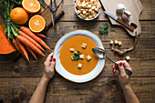 From above shot of unrecognizable person holding a bowl of orange carrot soup in a rustic wooden table with vegetables arrangements