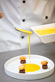 Crop cooker in uniform holding bowl and pouring yellow syrup from spoon into white plate with chocolate pastries in form of mushrooms