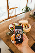 Hand of person using smartphone and taking photo of various dishes placed together on wooden table