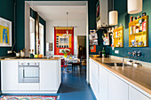 Kitchen counter with white cabinets, petrol-blue wall and dining room with colourful wall hanging in background