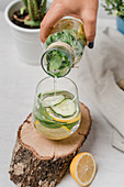 Faceless woman pouring tasty lemonade with slices of fresh lemon and cucumber in crystal glass on table