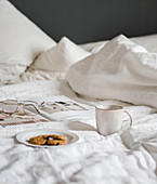 Plate of biscuits, mug, book and glasses on bed with white bed linen