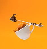 Falling white cup with splashing hot coffee and teaspoon with beans of coffee on yellow background