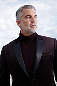 A grey-haired man on a terrace wearing a dark-red turtleneck jumper and a jacket