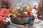 Schale mit herbstlichem Bastelmaterial: Hagebutten, Zweig, Äpfel, Herbstlaub, Maronen, Mais, Kränzchen aus Heide