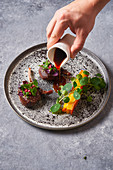Person adding sauce to meat medallions with herbs on plate on gray stucco table