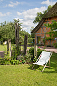 Liegestuhl in sommerlichem Garten, im Hintergrund Reetdachhaus