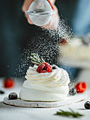 Woman sprinkles icing sugar on mini Pavlova cake decorated fresh berries and rosemary