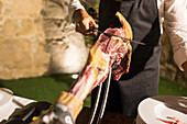 Crop unrecognizable person hand on apron cutting a whole dry-cured ham leg on a black background