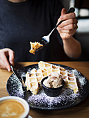 Unrecognizable person using form and knife to cut sweet waffles near bowl of ice cream and cup of coffee