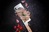 Huge knife with slices of fried meat placed near garlic cloves and tomatoes on black background