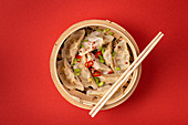 Traditional Chinese steamed dumplings in bamboo steamer with wooden chopsticks on red background, close up, top view
