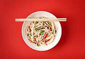 Asian noodles with vegetables, green peas and red pepper in white bowl with wooden chopsticks