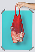 Hand carrying red string sack with glass containers of dairy against turquoise rectangle during eco friendly shopping