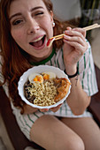 Cheerful redhead female laughing blinking eye and picking noodles from bowl of tasty ramen while sitting on couch at home