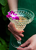 A woman in a green dress, standing outside by greenery holding a glass of prosecco with a purple orchid