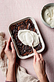 Hands spreading whipped coconut cream over a chocolate cake in a baking dish.