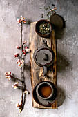 Tea drinking wabi sabi japanese style dark clay cup and teapot on wooden board with blooming cherry branches. Grey texture concrete background. Flat lay, space