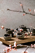 Tea drinking wabi sabi japanese style dark clay cups and teapot on wooden tea table with blooming cherry branches. Grey texture concrete background.