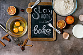 Kreidetafel mit Aufschrift 'Stay home and bake' zwischen Backzutaten