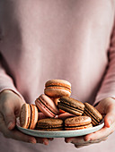 Heap of french macarons on pink plate in hands of woman dressed in pink clothes. Vertical. Copy space