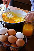 From above crop female using whisk to mix sugar with raw eggs near cup of citrus juice while cooking pastry on kitchen table