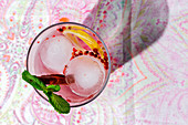 Gin tonic cocktail with pink tonic water, pink pepper, rosemary, mint, cinnamon, lemon and orange in the sunlight on a restaurant table