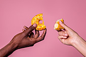Dunkle und helle Hand halten angebrochenes Donut vor rosa Hintergrund