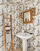 Bamboo mirror above pedestal sink and ladder in bathroom with patterned wallpaper