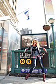 A young blonde woman wearing a down jacket, an owl t-shirt, a checked shirt and jeans standing in front of a Metro sign