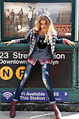Junge blonde Frau in Daunenjacke, Shirt mit Eulenprint, Karohemd und Jeans vor U-Bahn Schild