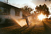 Child running through lawn sprinkler as sun sets