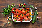 Various types of tomatoes in an enamel vessel next to cucumber, herbs and garlic