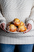 Crop female in white sweater and jeans holding plate with homemade tasty fresh buns with white sprinkles