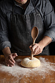 Using big wooden spoon for making hole in dough while forming artisan round bread loaf at wooden table