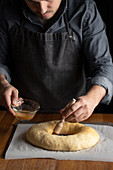 Crop male chef in black apron greasing unbaked round bread with egg yolk while standing at wooden table