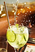 Splashing flow of alcoholic mojito cocktail in crystal glass with lime slices