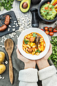 Bowl of white bean soup with chopped pork sausages and vegetables above table with ingredients and guacamole in mortar
