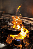 Chef tossing frying shrimps in flambe above gas stove with burning flame while cooking in restaurant
