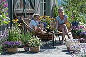 Mutter und Sohn sitzen auf insektenfreundlicher Terrasse mit blühendem Basilikum, Scheinsonnenhut, Malve, Fächerblume 'Violet Blue', Zweizahn 'Bee White', Rosmarin