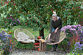 Small seating group in front of crabapple tree, woman puts blanket on armchair, autumn asters, chilli 'Bolivian Rainbow' and red grass in the flowerbed