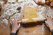 Anonymous female in black dress rolling out dough with wooden rolling pin on table with flour and various kitchen utensils while making pastry for pasta