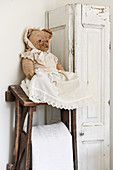 An old teddy bear wearing a white dress next to a white shabby chic-style cupboard