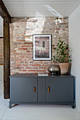 Entrance area with white painted wooden floor and brick wall, in front of it a gray painted lowboard