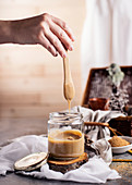 Hand holding teaspoon over transparent glass jar of tasty caramel