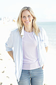 A blonde woman by the sea wearing a purple t-shirt and a blue-and-white striped shirt