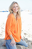 A blonde woman by the sea wearing an orange tunic and jeans