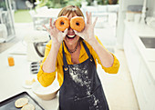 Mature woman covering eyes with donuts in kitchen