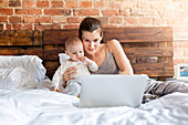 Mother with baby daughter using laptop on bed