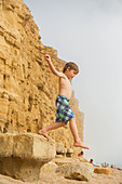 Boy in swim trunks jumping on beach rock